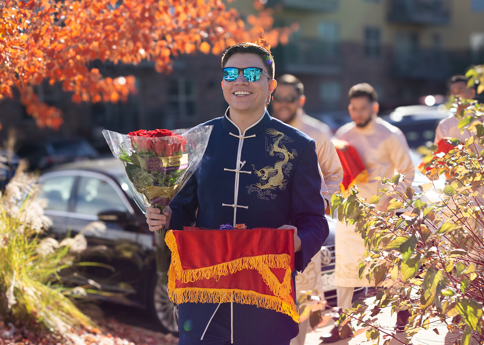Groom wearing dark blue chinese outfit and blue sunglasses carrying gift tray to tea ceremony