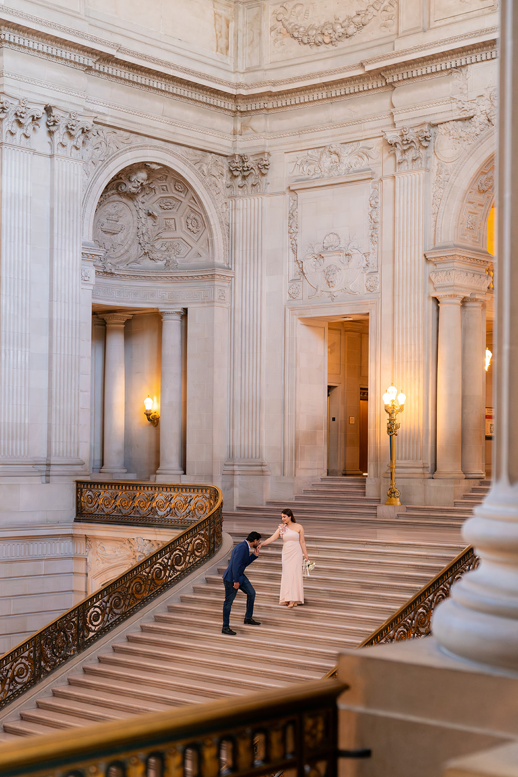image of male and female couple on grand steps at san francisco city hall. man is slightly bowed to kiss the woman on her hand