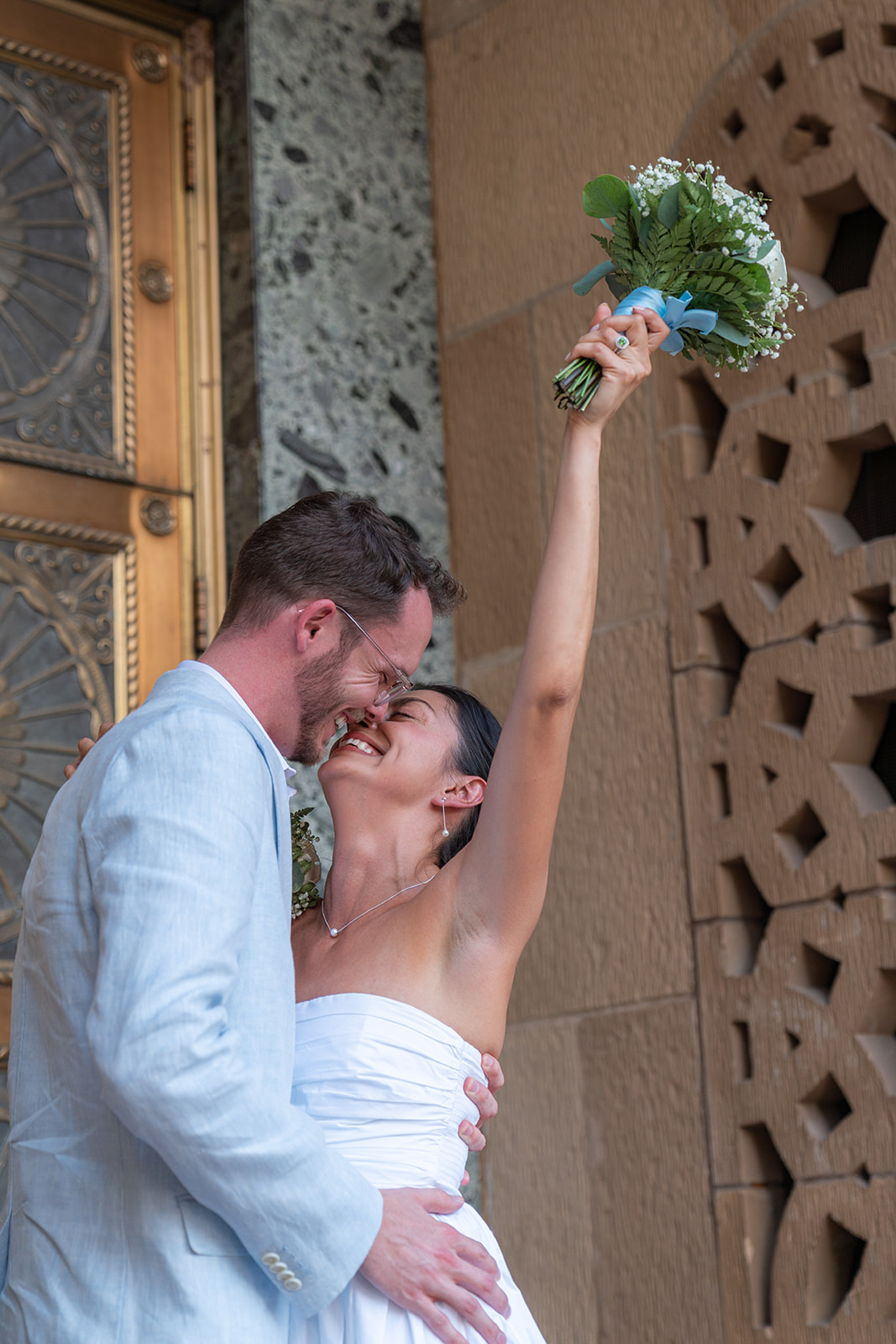 Couple smiling and kissing. Groom is wearing a light blue suit and bride is wearing a strapless white dress. The bride's arm is extended into the air and she is holding a bouquet of white roses. They are at the Phoenix City Hall after getting married at the Phoenix Courthouse