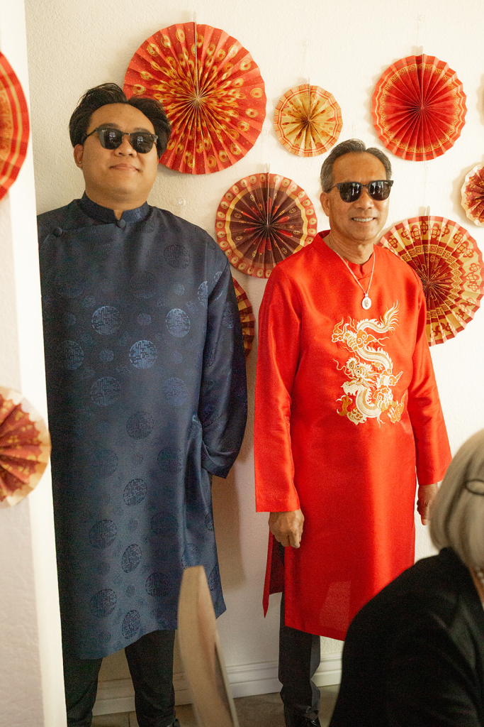 two men wearing sunglasses standing next to each other. one is wearing a dark blue vietnamese ao dai and the other is wearing a red one with a gold dragon