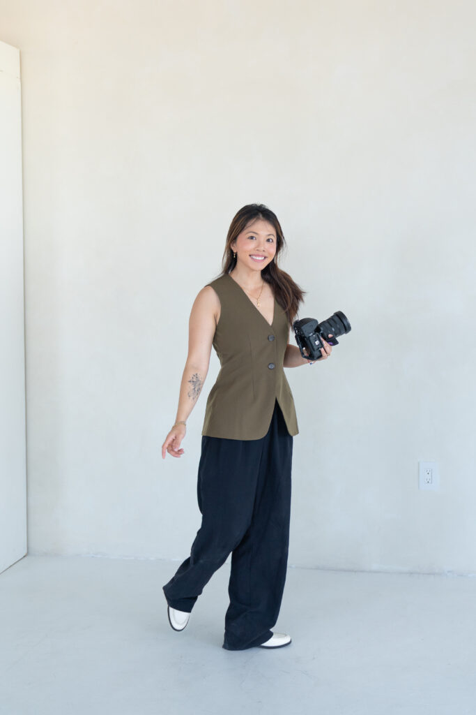 A woman, Tran Tran, walking with a camera in her right hand. She is wearing an olive-colored vest with dark blue trousers and white platform shoes