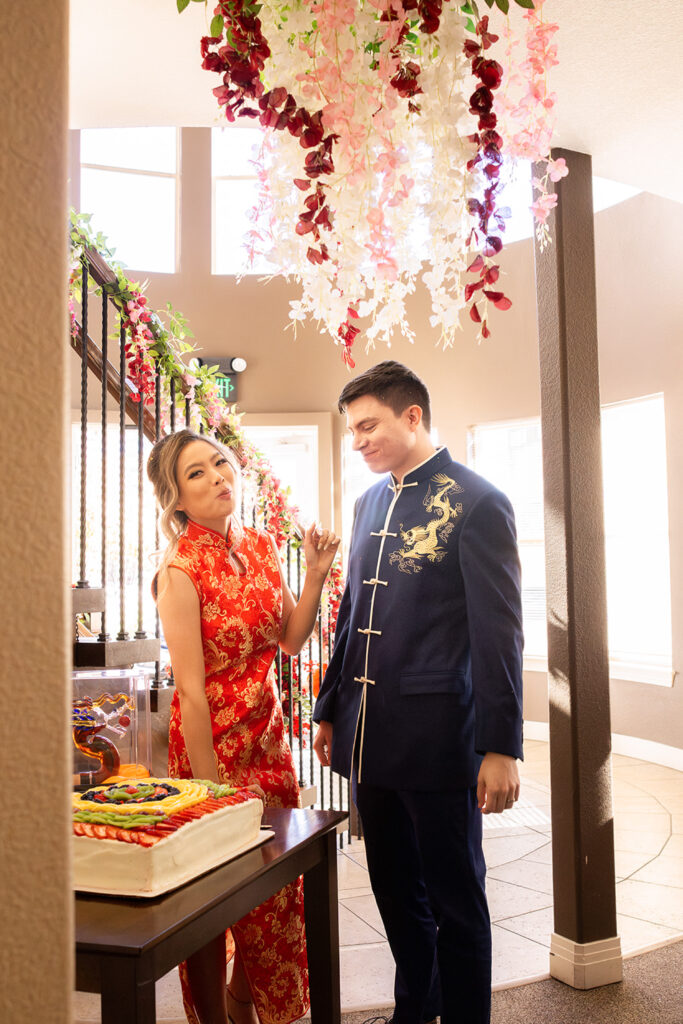 Asian bride wearing red qipao and smiling at white groom in blue tang suit while cutting wedding cake