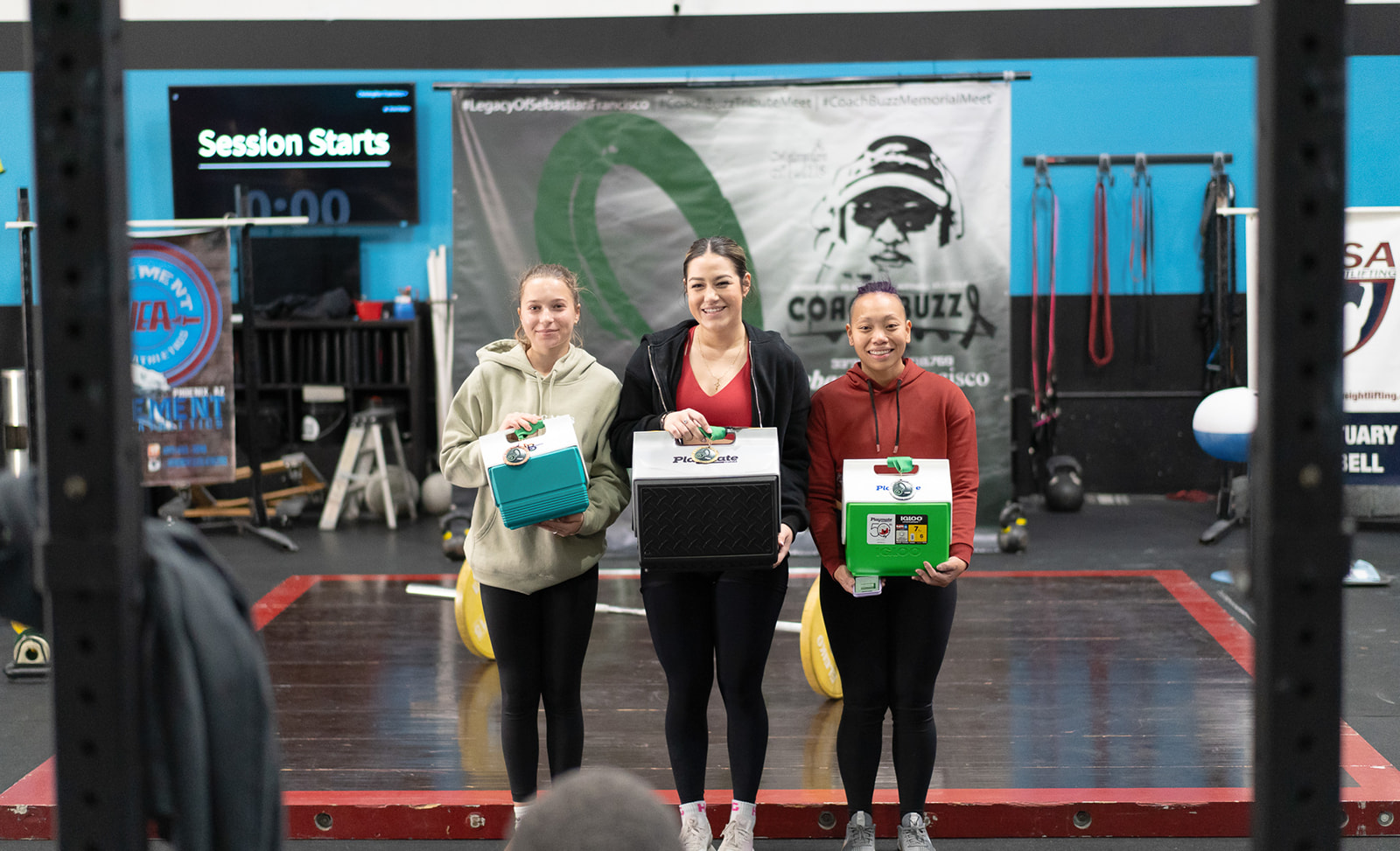 three women standing on a podium after a weightlifting meet. They are holding coolers