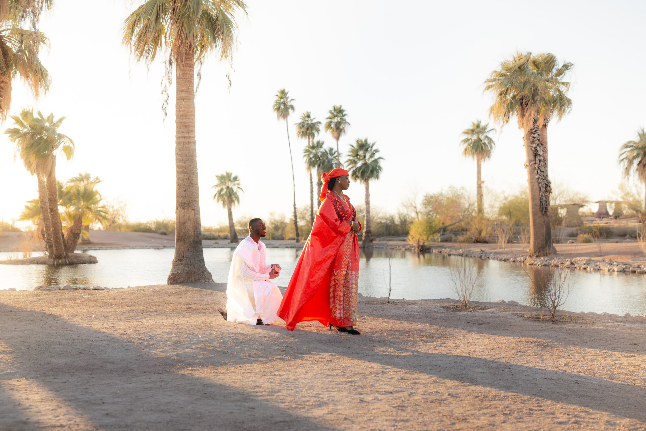 Surprise proposal moment where the woman does not know she will be proposed to. The man is behind her on one knee. They are surrounded by a body of water and tall palm trees.