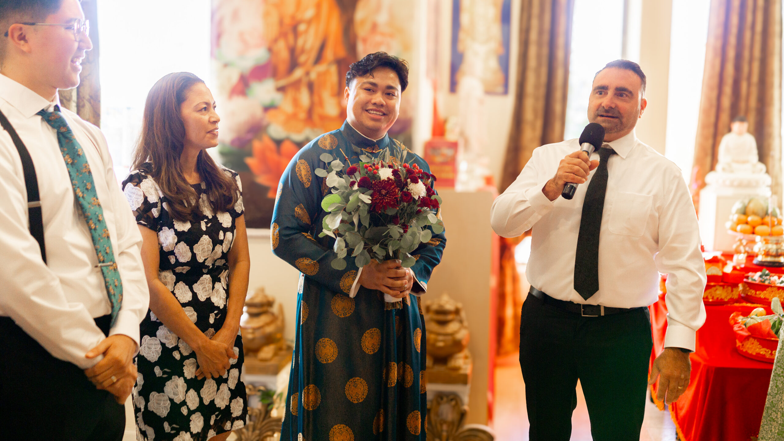 three people at a tea ceremony. a man is talking into a microphone, while the groom is smiling holding a bouquet of flowers