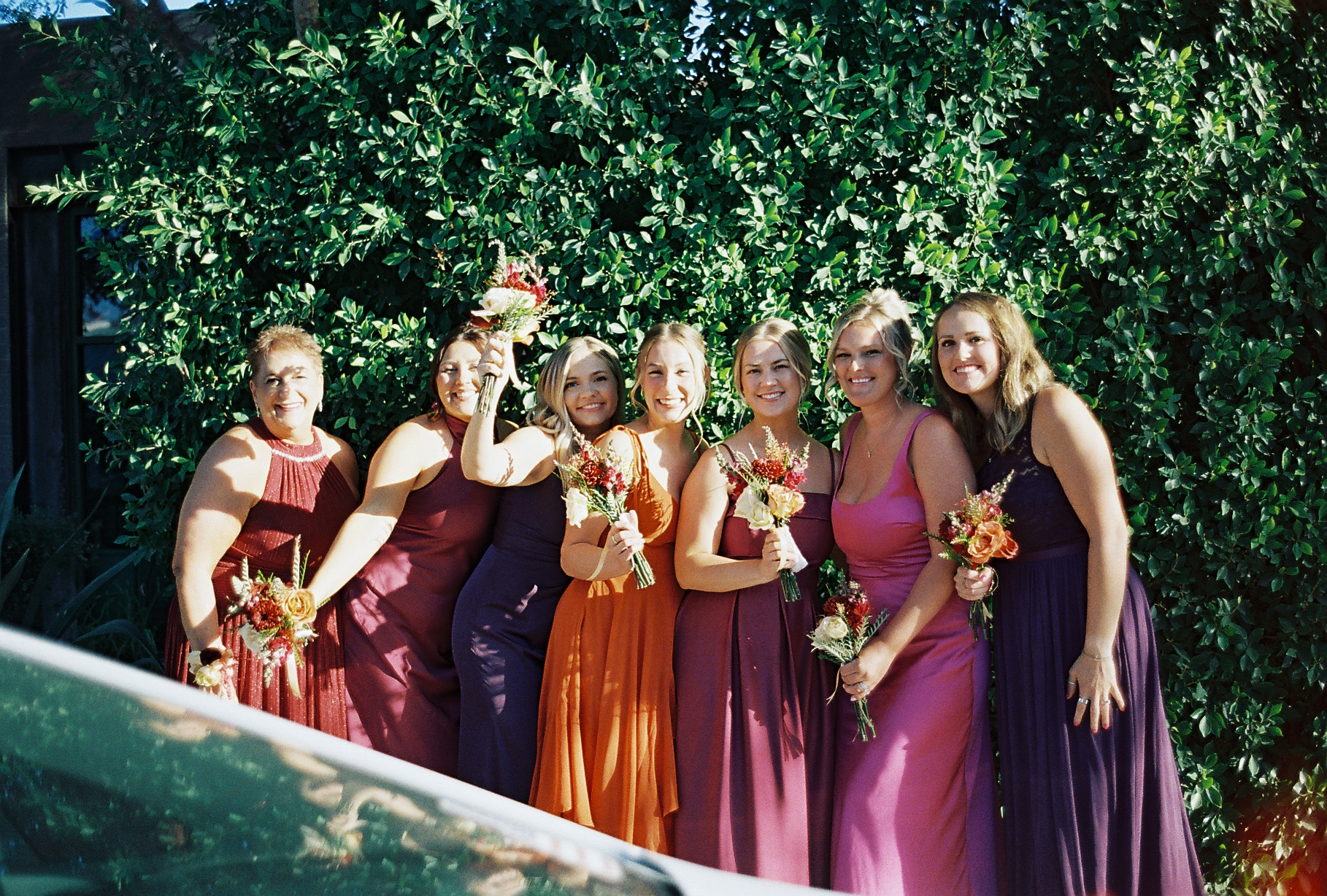 Bridesmaids wearing different-colored dresses with bouquets in front of a green leaf wall