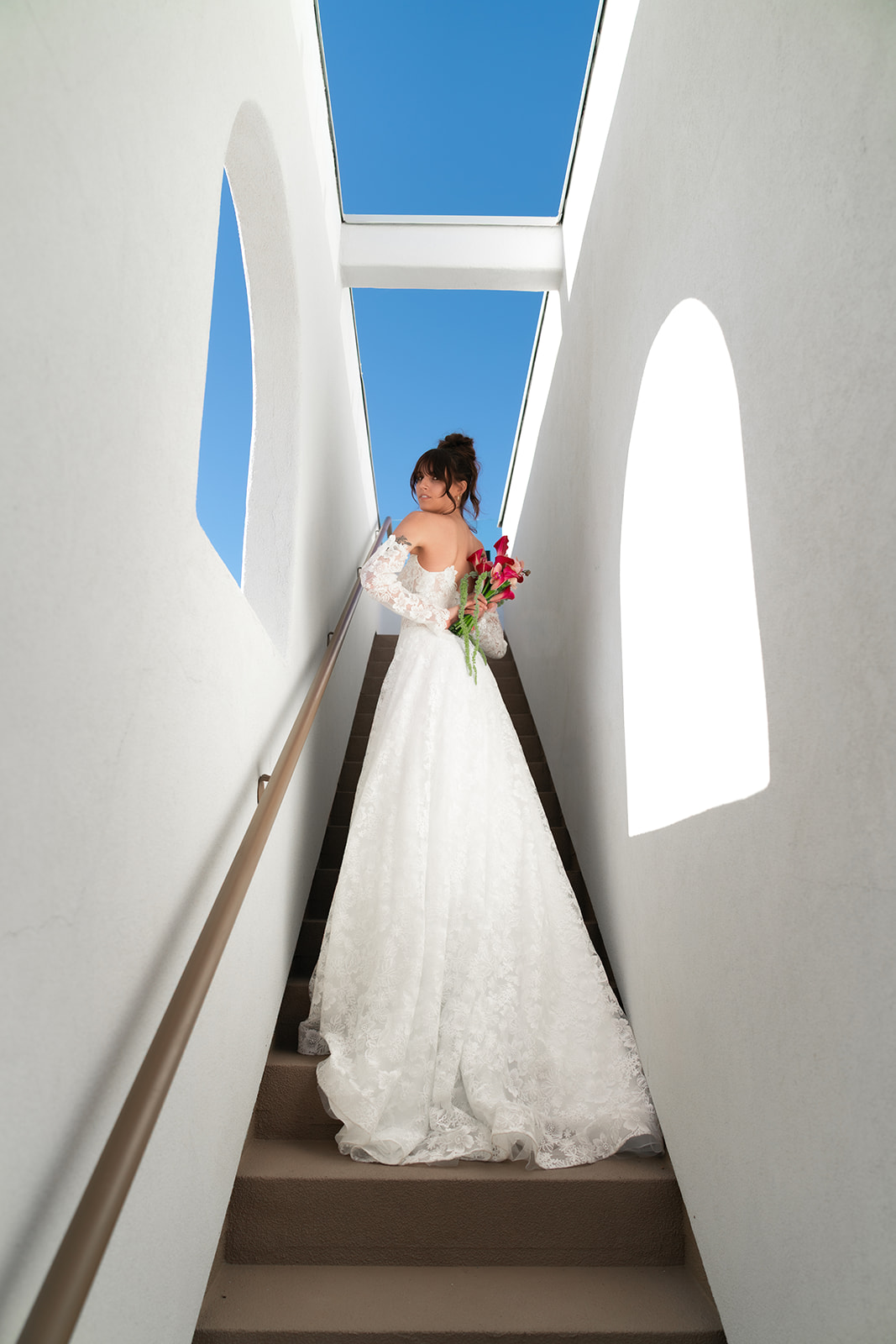Bride standing on stairs with bright blue sky in the background. She is holding her bouquet behind her back